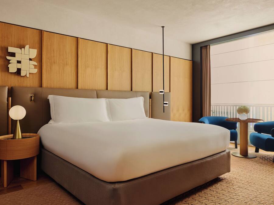 Ein modernes Hotelzimmer mit einem großen Bett, einem hölzernen Kopfteil, einem Nachttisch mit Lampe und Blick auf den Balkon.