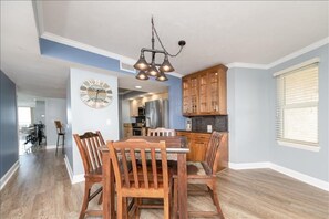 A dining area with a wooden table and chairs, a wall clock, and a kitchen in the background.
