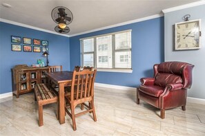 A dining area with a wooden table and chairs, a red leather chair, a wall clock, and a window with blinds.