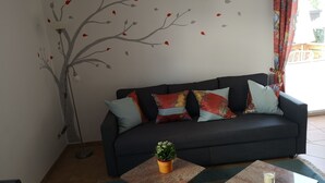 A dark-colored sofa with multiple cushions, a small potted plant, and two square tables with candles. A wall mural of a tree with leaves is visible. - Vacation apartment Hermann (Bovenden)