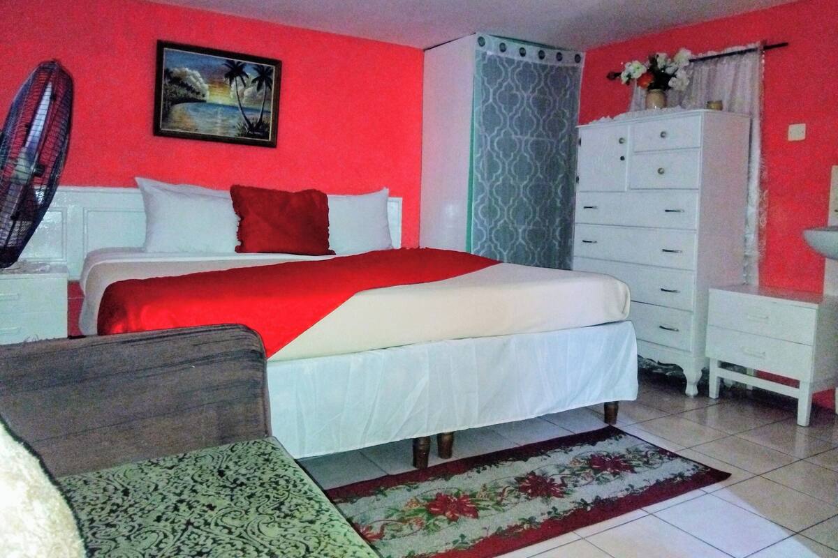 Un dormitorio con cama, cómoda, mesita de noche, ventilador y un cuadro en la pared.