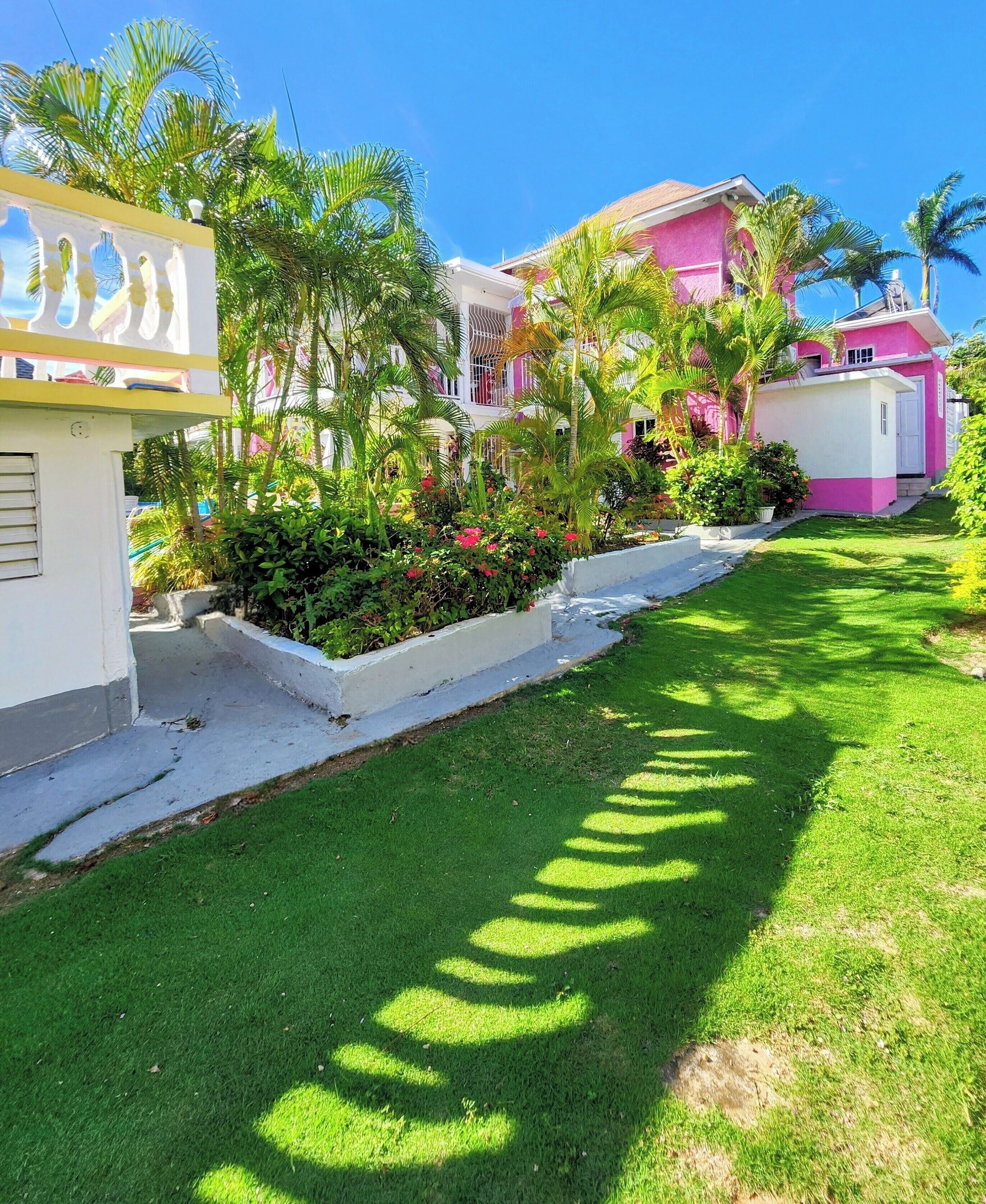 Un barrio residencial con un césped bien cuidado, un edificio rosa y abundante vegetación.