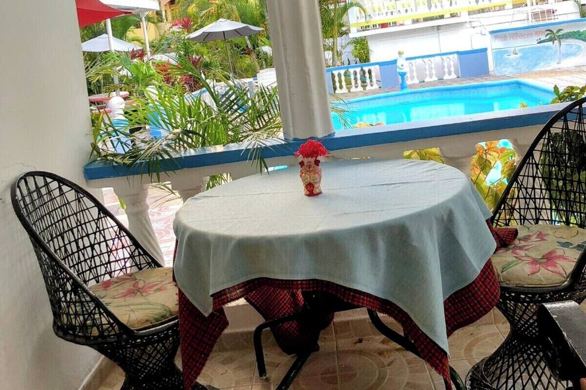 Un patio al aire libre con una mesa redonda y dos sillas, un jarrón decorativo y vistas a una piscina y follaje.