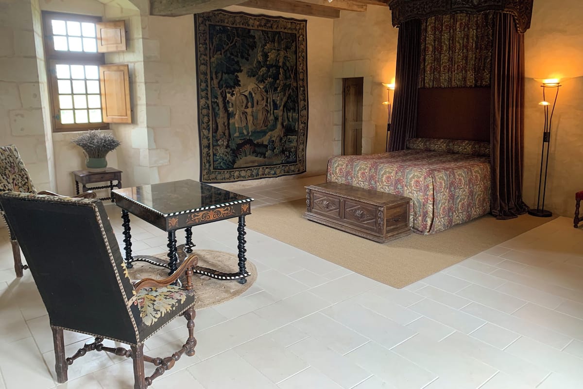 A bedroom with a four-poster bed, a large tapestry, a small table, and a chair.