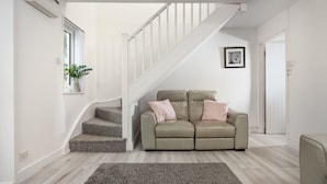 A living room with a beige sofa, a grey rug, a staircase, and a window with a plant. - Sunshine Cottage -  a cottage that sleeps 4 guests  in 2 bedrooms (Padstow)