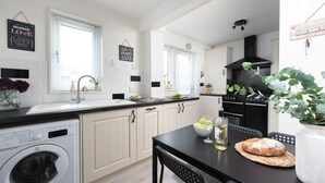 A modern kitchen with a built-in washing machine, a dining area with a table and chairs, and a black stove. - Sunshine Cottage -  a cottage that sleeps 4 guests  in 2 bedrooms (Padstow)