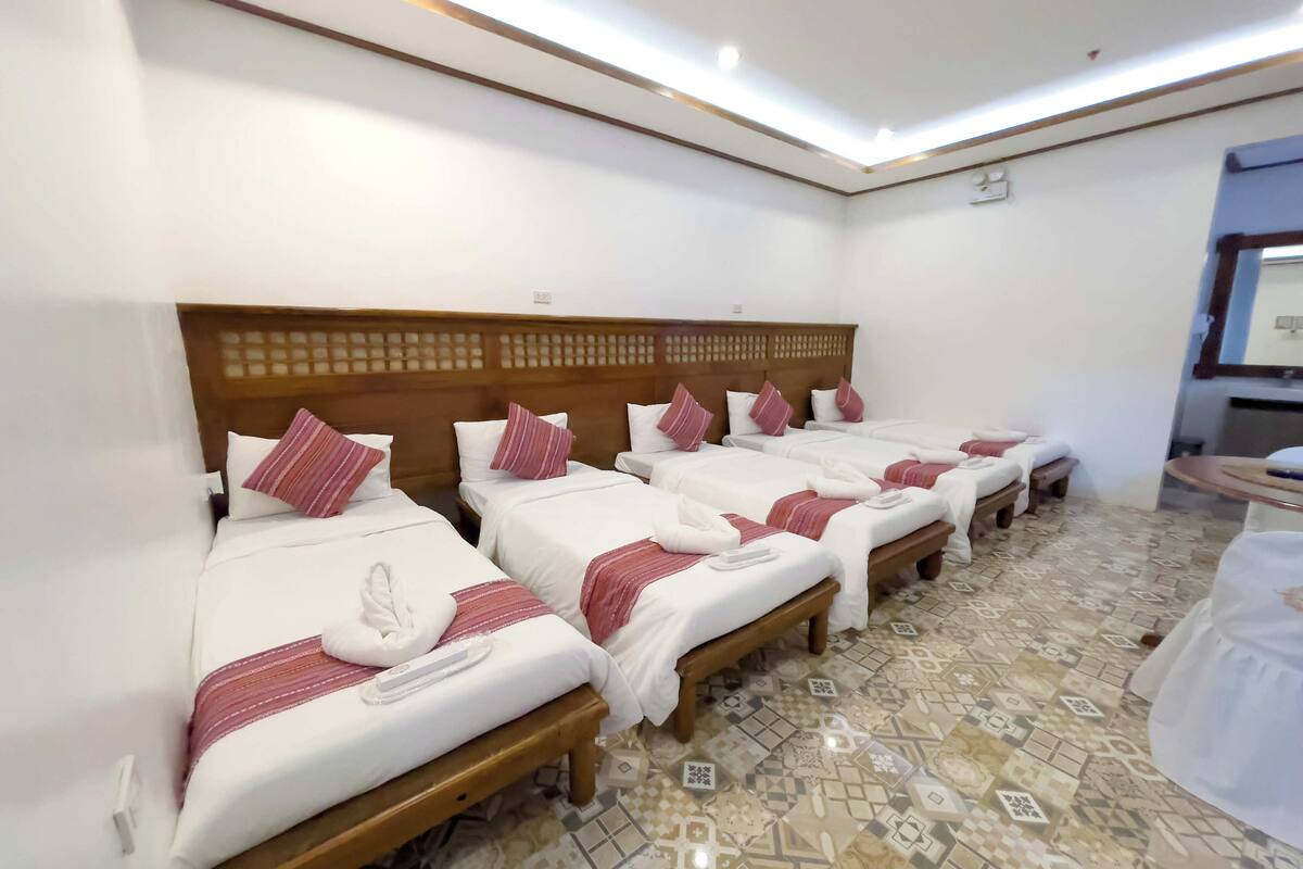 A hotel room with four neatly arranged beds, each with a wooden headboard, white bedding, and red striped pillows. The room has a patterned tile floor and a small table with a chair.