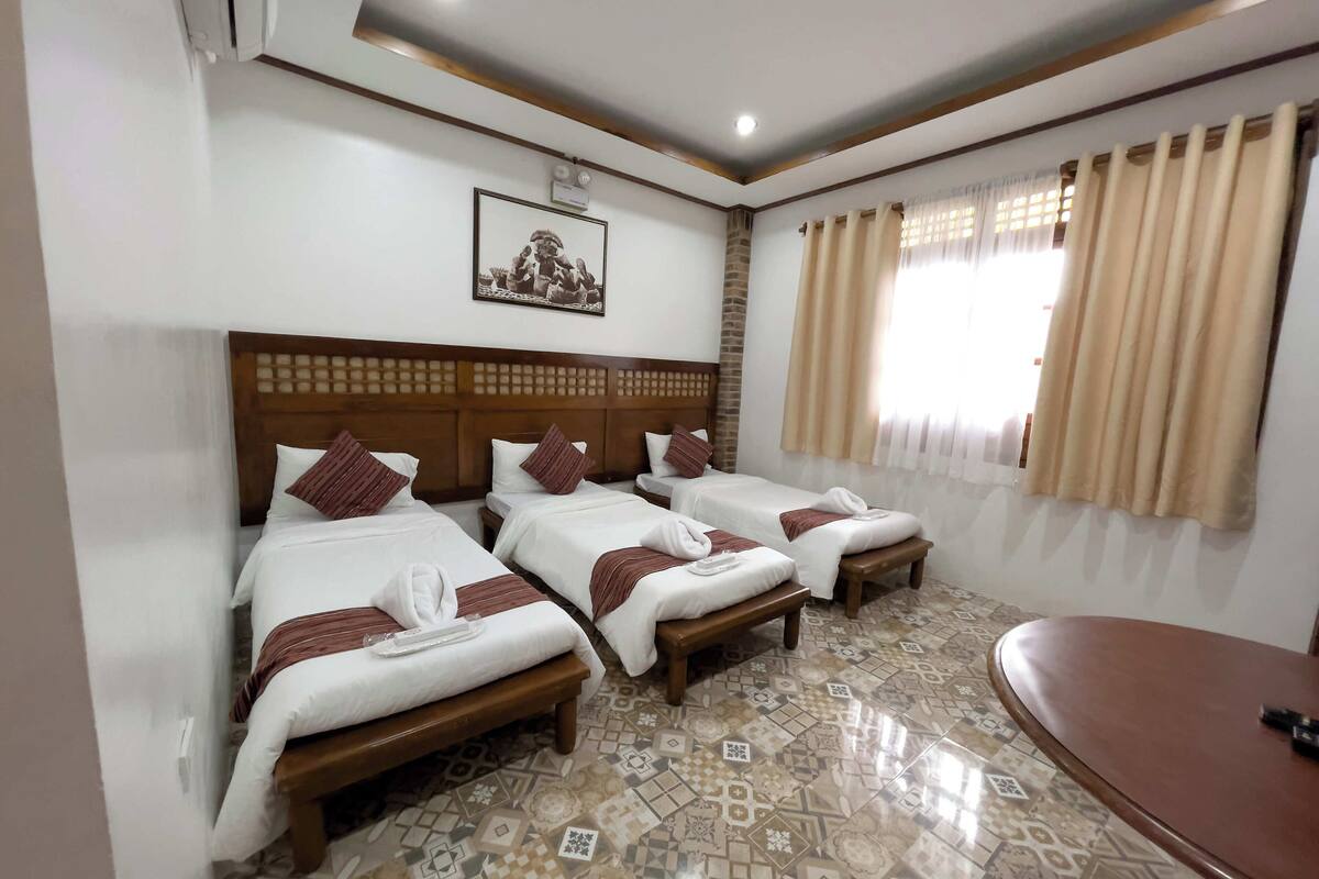 A hotel room with three beds, a wooden headboard, a patterned floor, and a window with curtains.