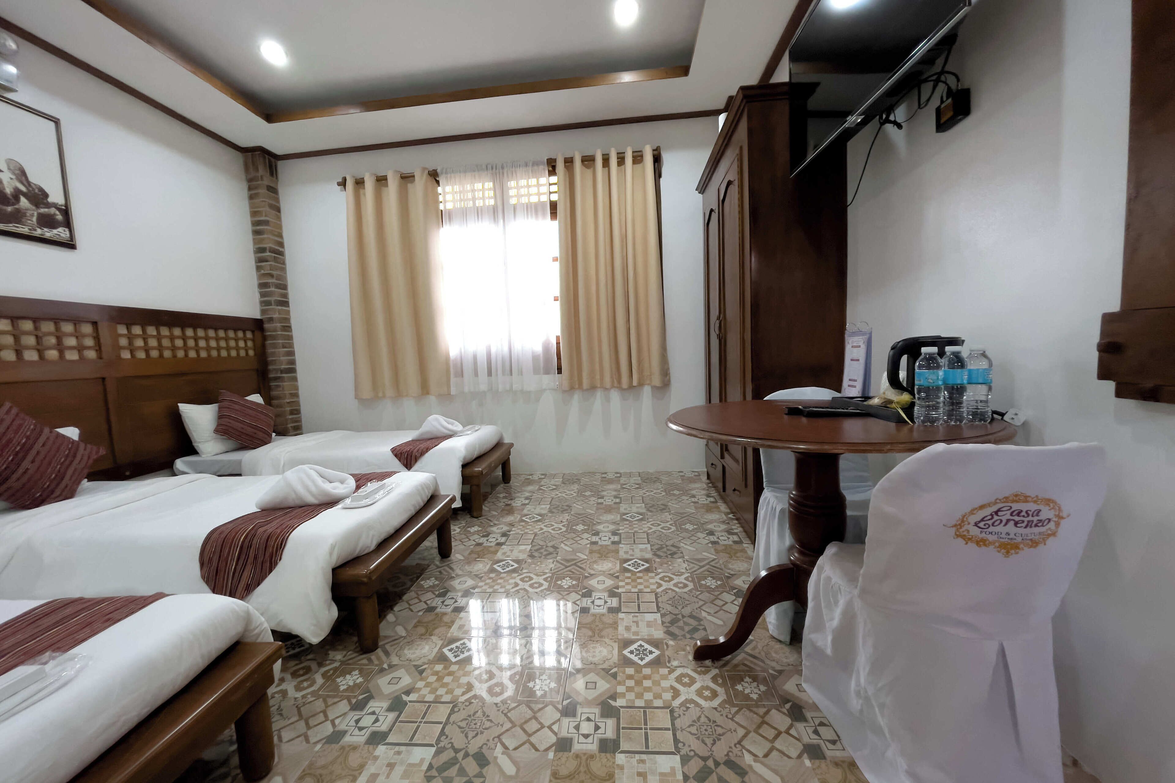 Habitación de hotel con tres camas, cabecera de madera, suelo estampado, un escritorio con silla y una ventana con cortinas.