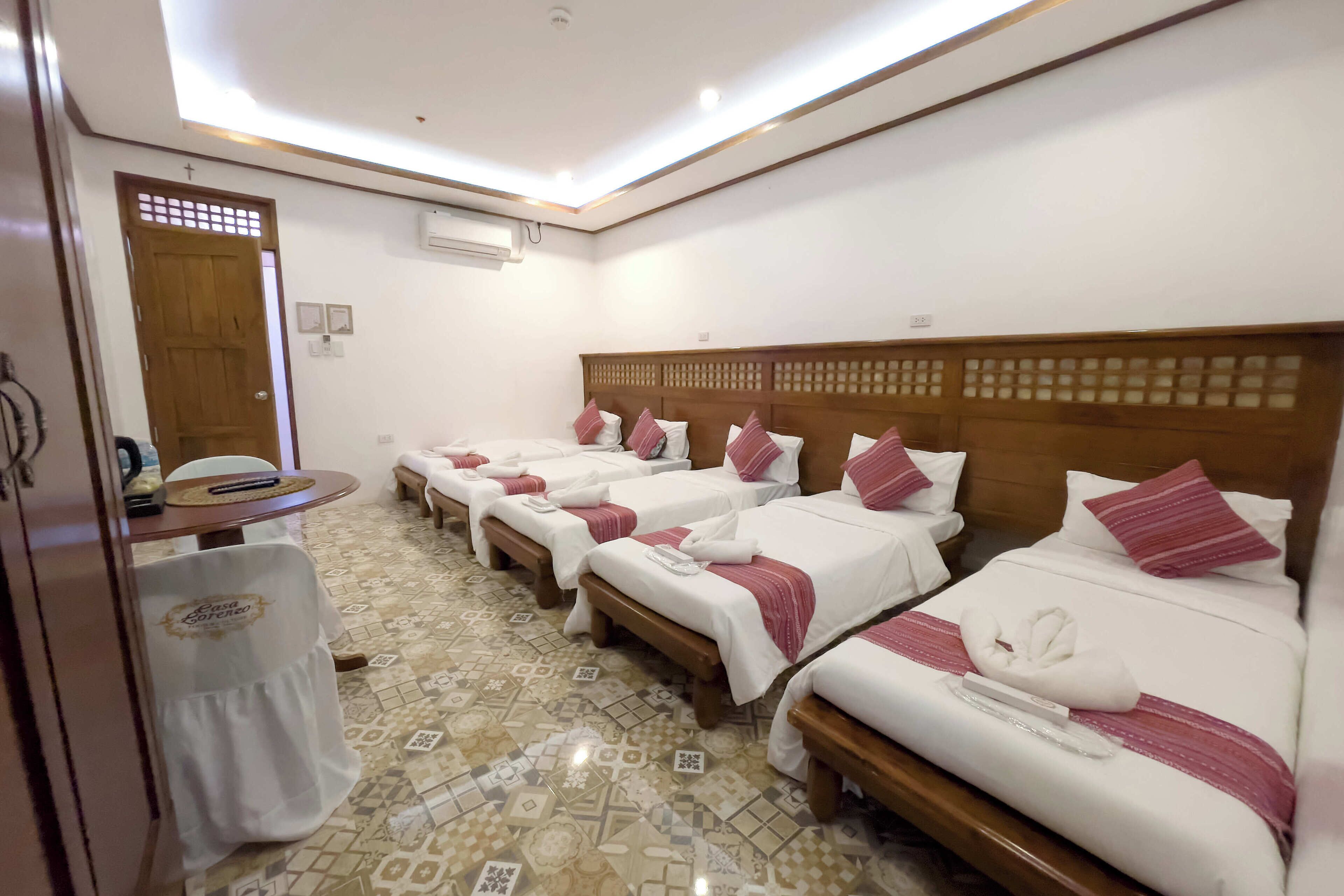 Ein Hotelzimmer mit vier Betten, jeweils mit weißer Bettwäsche und pinkfarbenen Akzenten, einem Holzkopfende und einem gemusterten Boden.
