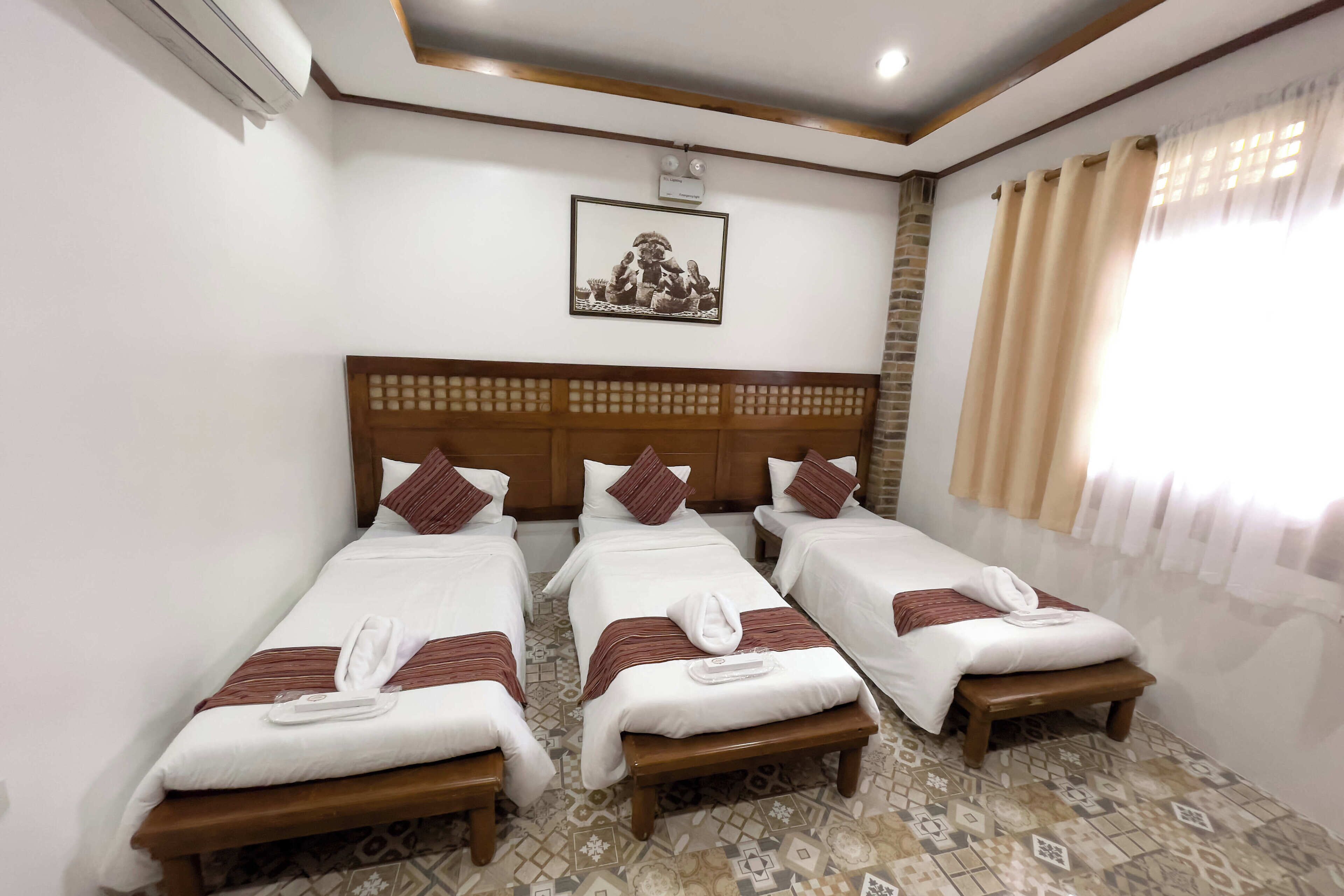 Habitación de hotel con tres camas individuales, cada una con cabecero de madera, ropa de cama blanca y una manta marrón. Al pie de cada cama hay un taburete de madera pequeño. La habitación tiene baldosas de suelo con diseño y una ventana grande con cortinas color beige.