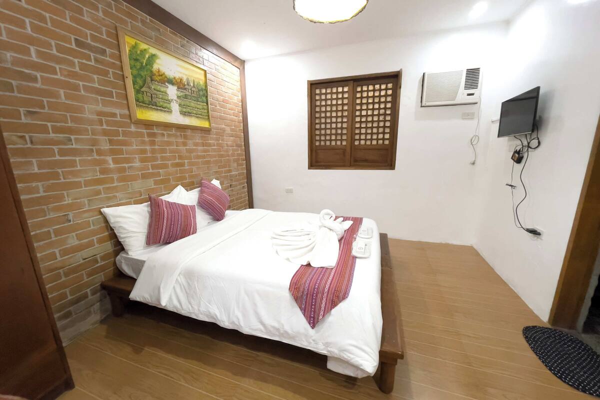 A neatly made bed with white linens and a red and white striped blanket, a wooden bench beside it, a brick wall with a framed painting, a window with wooden lattice, and a wall-mounted TV.