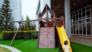 A playground with a slide and climbing wall, surrounded by a well-maintained lawn and tall buildings in the background. - Deluxe Family Room in Tagaytay With Taal Lake View (Tagaytay)
