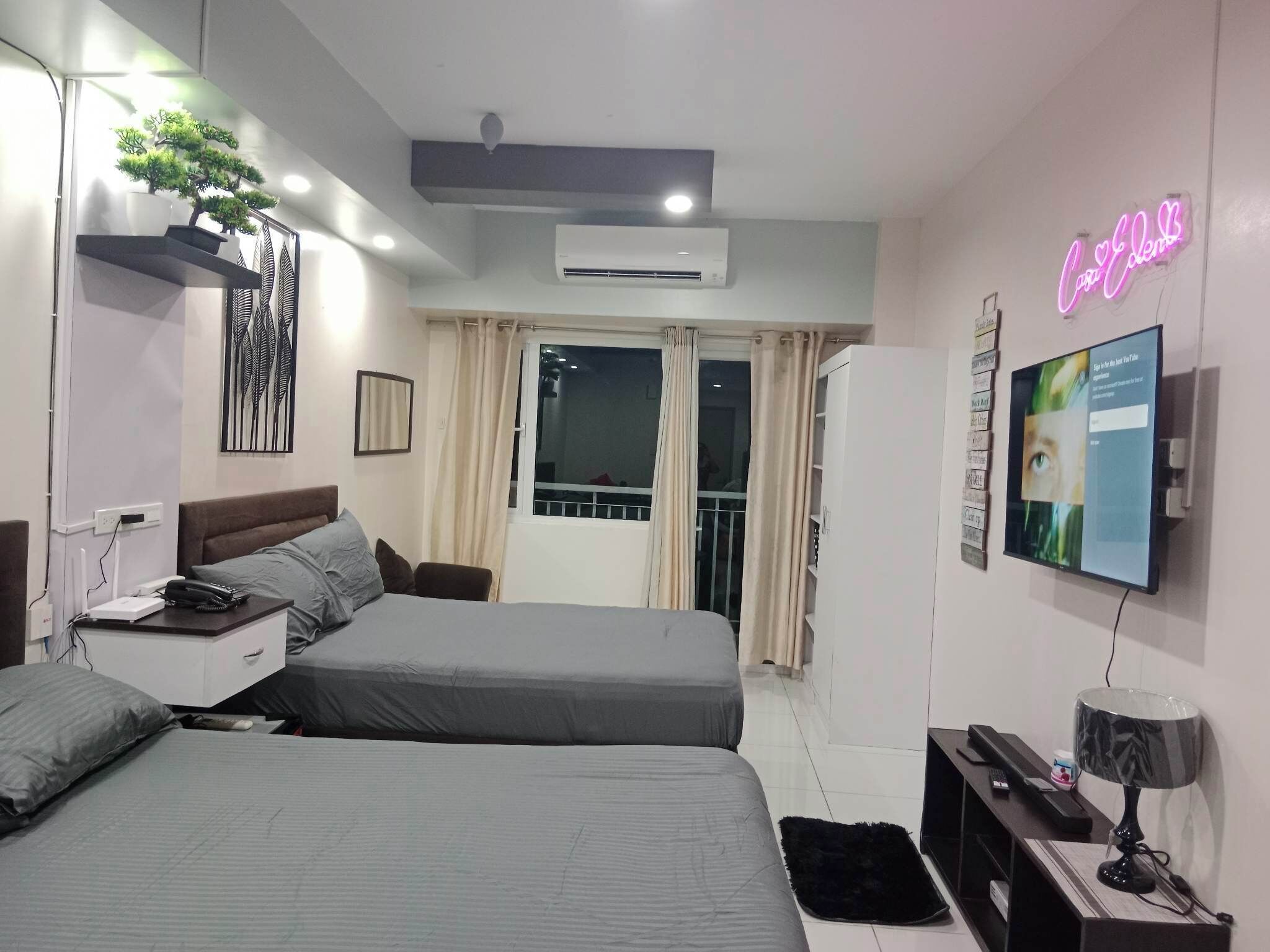 A modern hotel room with two beds, a flat-screen TV, a neon sign, and a balcony with curtains.