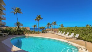 A clear blue pool with lounge chairs and palm trees, surrounded by a building with balconies.