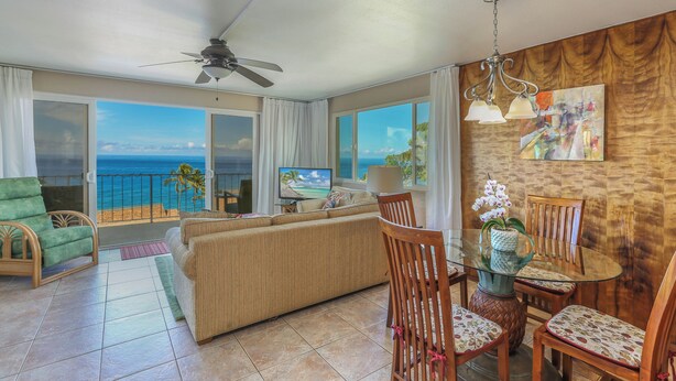 A spacious living area with a balcony overlooking the ocean, a comfortable sofa, a dining table with chairs, and a ceiling fan.
