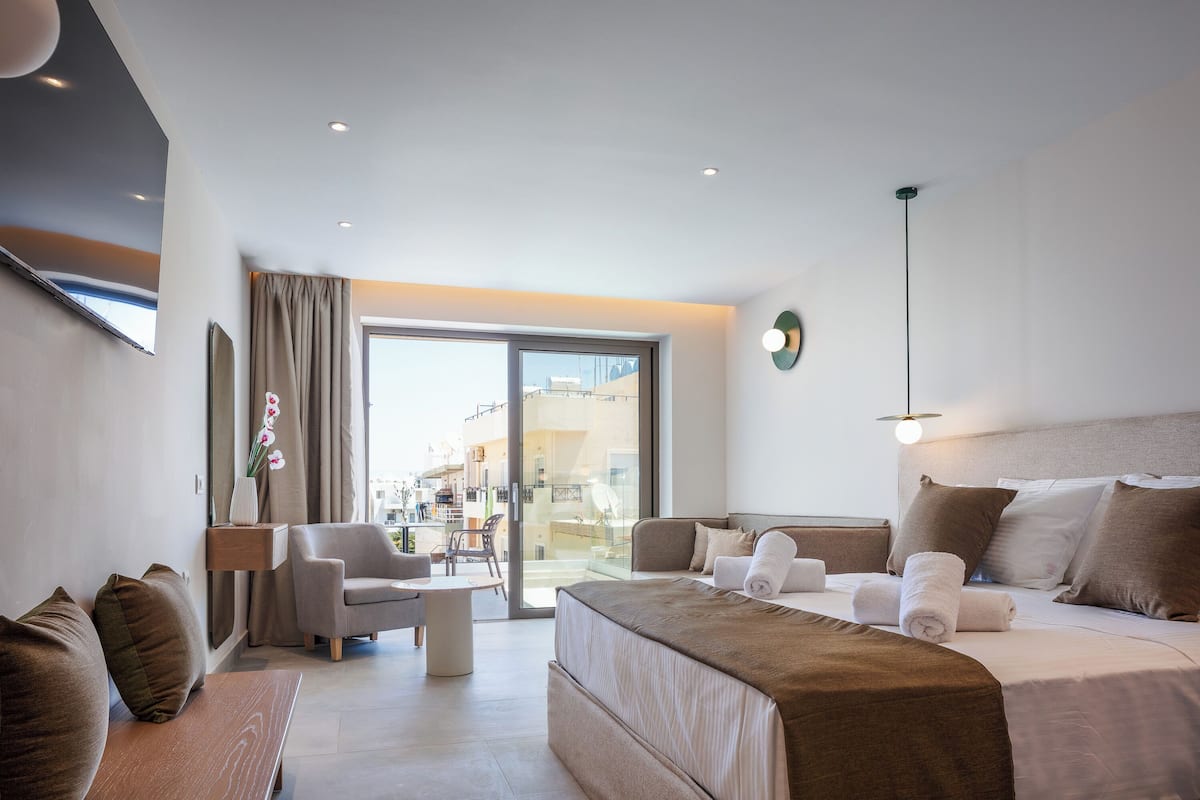 A modern hotel room with a large bed, a sofa, a desk, and a view of the city through a large window.