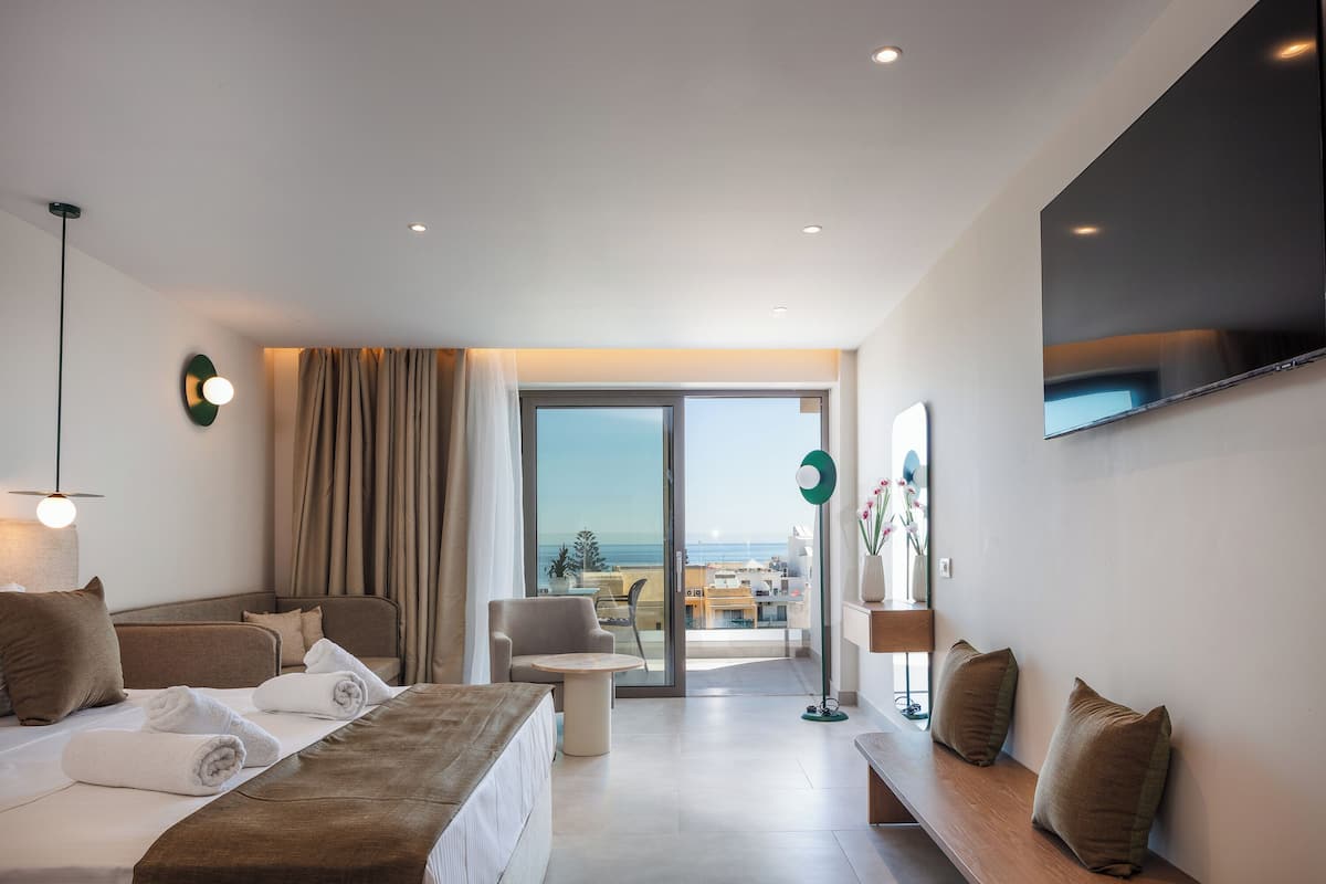 A modern hotel room with a bed, a sofa, a round table, and a TV mounted on the wall.