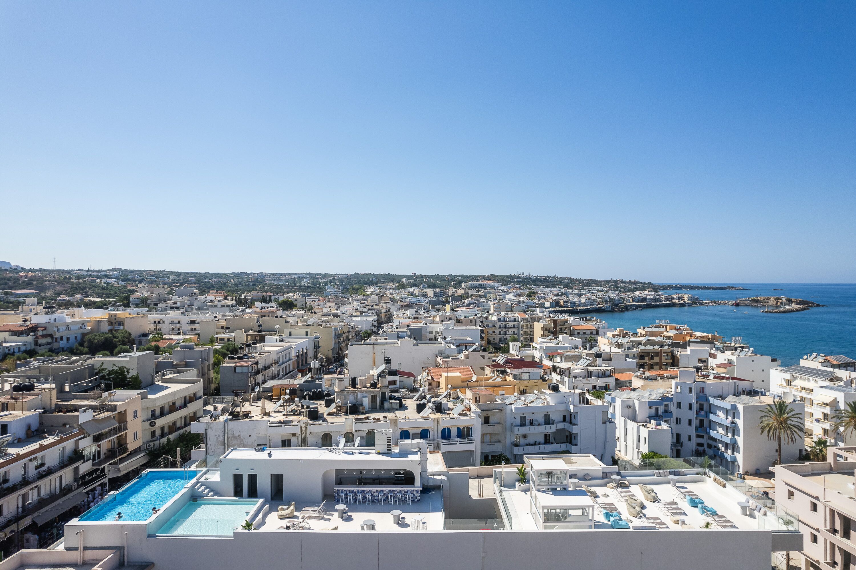 A coastal cityscape with a clear view of the sea, numerous buildings, and a rooftop pool area.