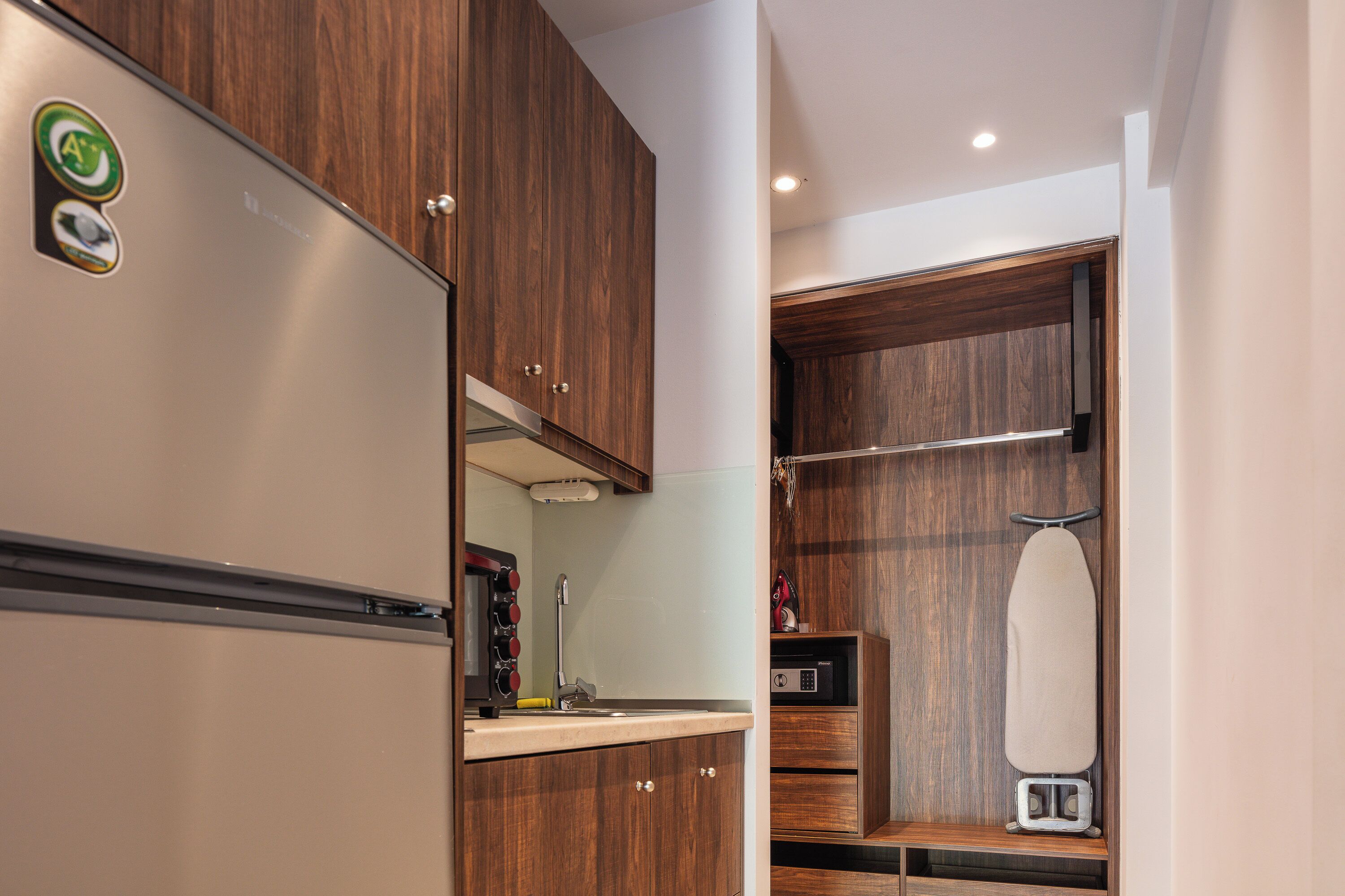 A compact kitchen with wooden cabinets, a stainless steel refrigerator, a microwave, and an ironing board.