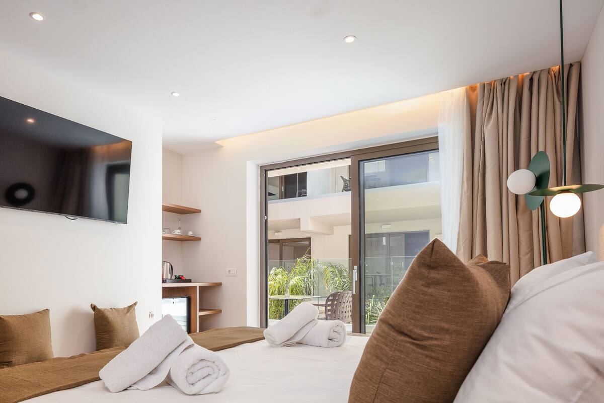 A modern hotel room with a bed, a TV, a balcony with a view, and a small kitchenette.