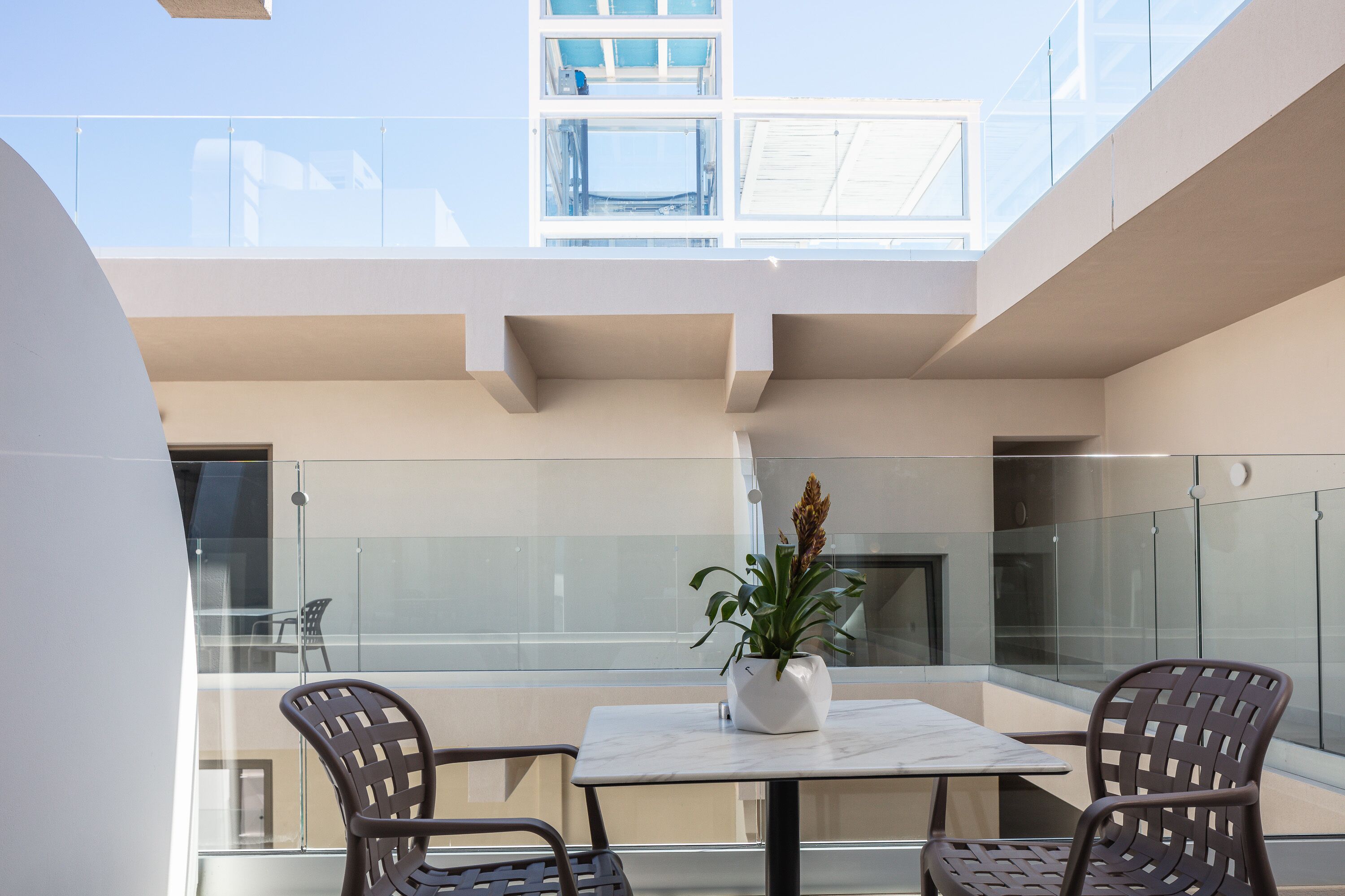 Un balcone moderno all'aperto con ringhiera in vetro, un tavolo bianco, due poltrone in vimini e una pianta in vaso.