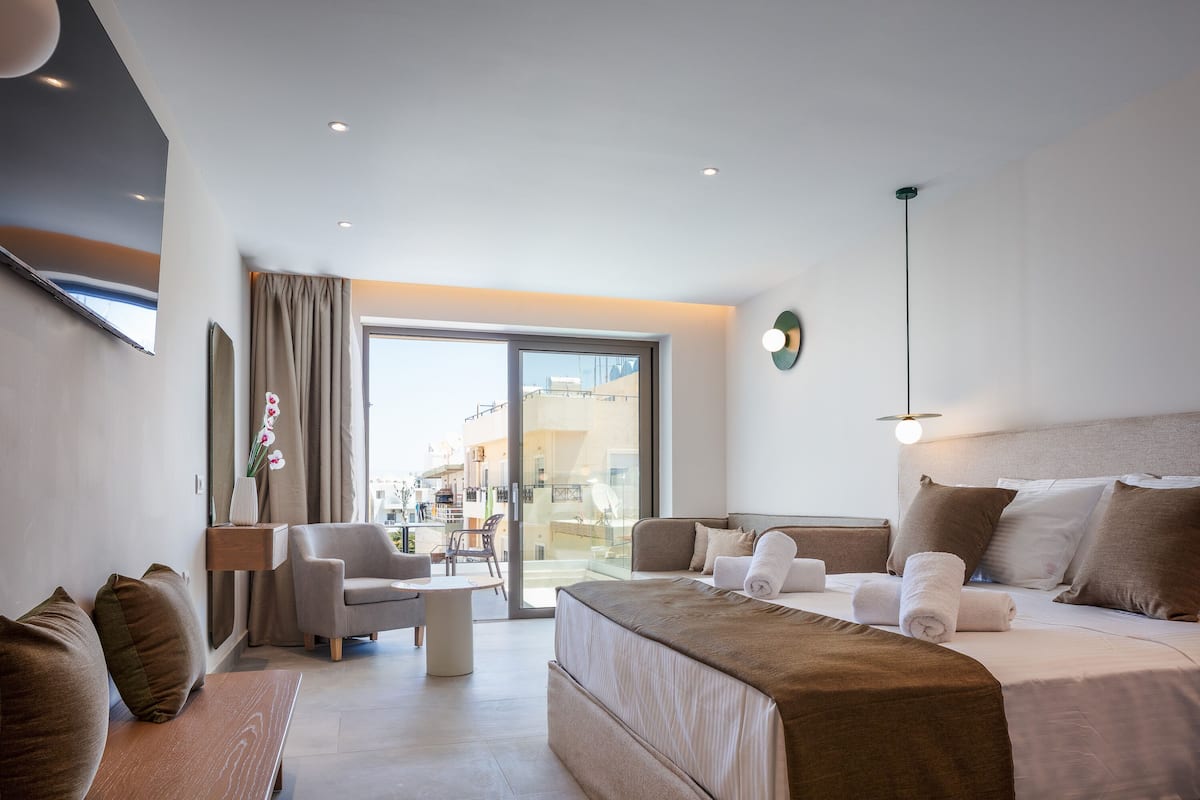 A modern hotel room with a large bed, a sofa, a desk, and a view of the city through a large window.