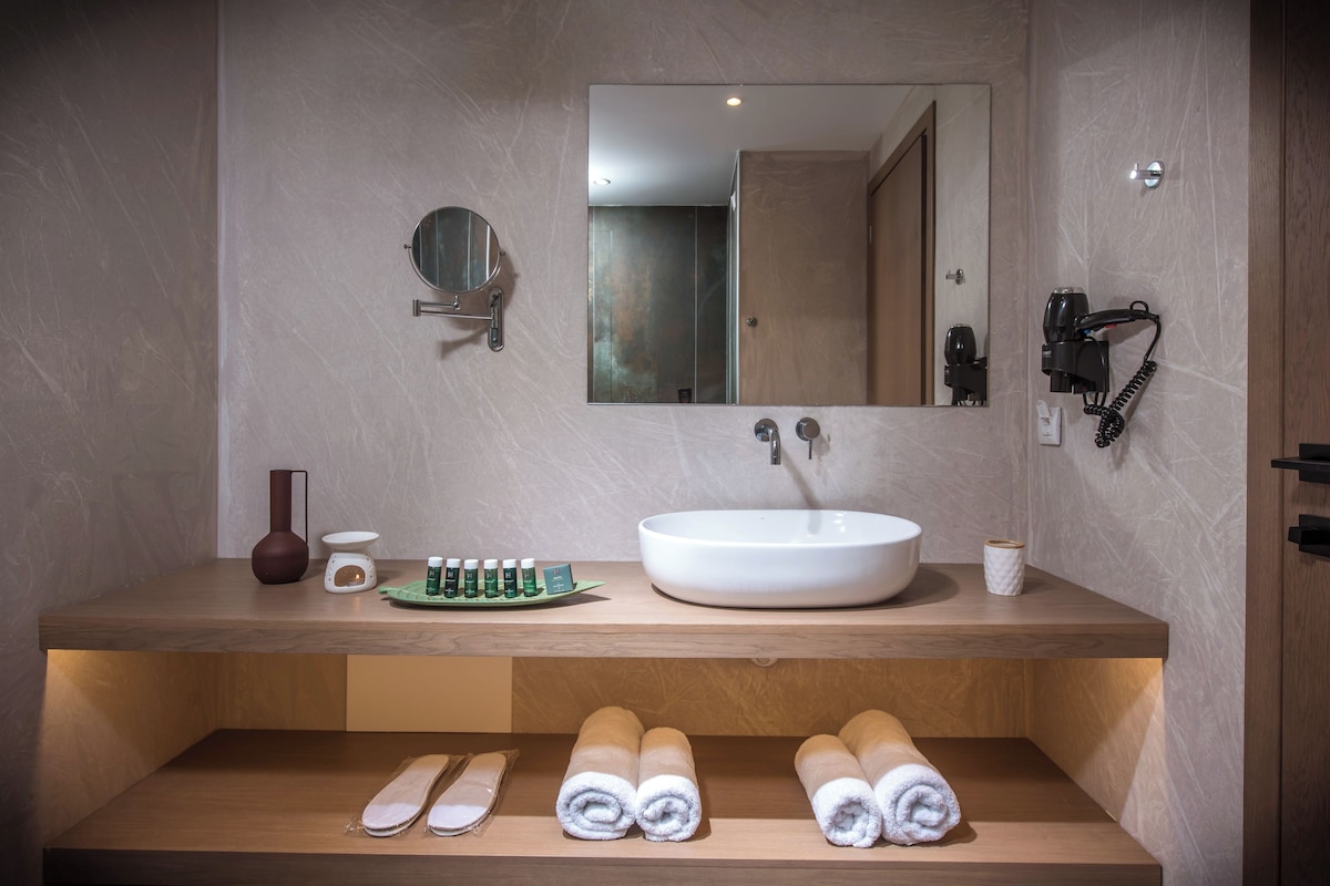 Un baño moderno con un espejo grande, un florero redondo, un lavamanos y toallas enrolladas con cuidado.