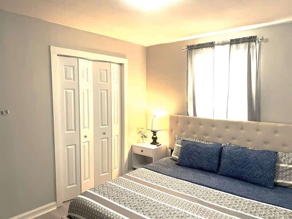 Un dormitorio con cama, mesita de noche, lámpara, ventana con cortinas y puerta.