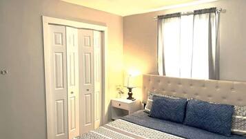 Un dormitorio con cama, mesita de noche, lámpara, ventana con cortinas y puerta.