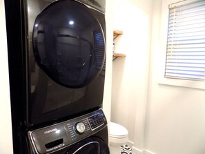 A black washing machine with a control panel, a white toilet, and a window with blinds in a bathroom.