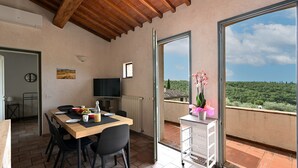 Un coin repas avec une table dressée pour deux personnes, un téléviseur et une vue sur un paysage verdoyant par le biais d’une grande fenêtre.