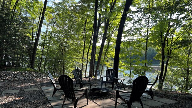 Zona de estar junto al lago con sillas Adirondack y una chimenea, rodeada de árboles y vistas al agua.