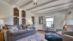 A living room with a grey sofa, a patterned rug, and a fireplace. There are framed pictures on the wall and a window with a view of the outdoors.