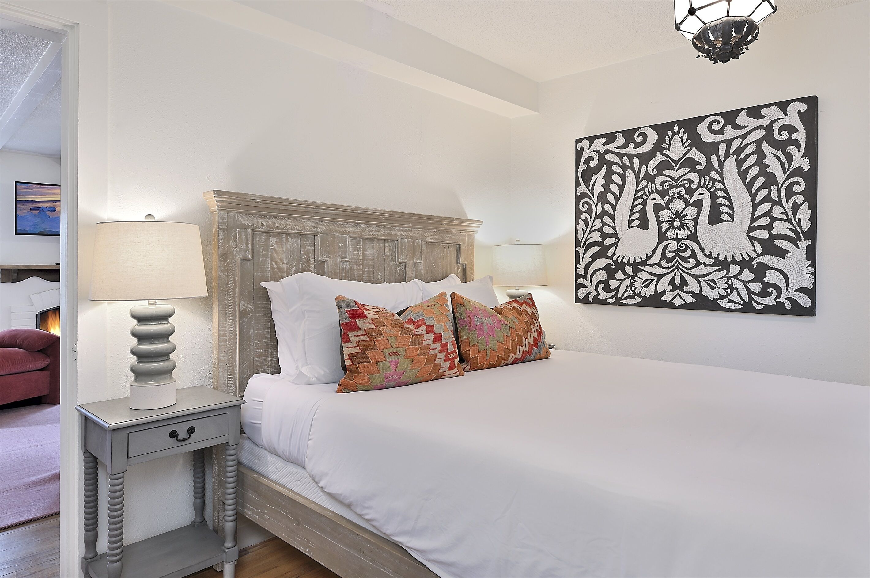 Una cama bien hecha con ropa blanca y almohadas estampadas, cabecera de madera, mesita de noche con lámpara y una obra de arte colgada en la pared.