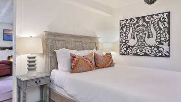 Una cama bien hecha con ropa blanca y almohadas estampadas, cabecera de madera, mesita de noche con lámpara y una obra de arte colgada en la pared.