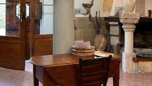 Zona pranzo con tavolo e sedie in legno, colonna in pietra con scultura a busto, e interni rustici con travi in legno e pavimento in piastrelle.