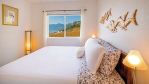A bedroom with a large bed, a window with a view of mountains, a bedside lamp, and wall art.