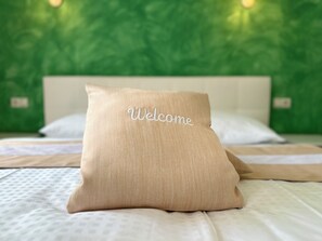 Un lit avec un oreiller beige sur lequel est inscrit le mot « Bienvenue », et un mur vert à texture visible en arrière-plan.