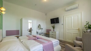 A hotel room with a bed, a desk, a chair, a wardrobe, a television, and an air conditioning unit. - Albizia-Apartments (Baden)