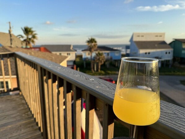 A glass of orange juice on a wooden deck with a view of houses and palm trees in the background. - Steps to the Beach | Surfside Home w/ Private Beach Access - pets welcome! (Surfside)