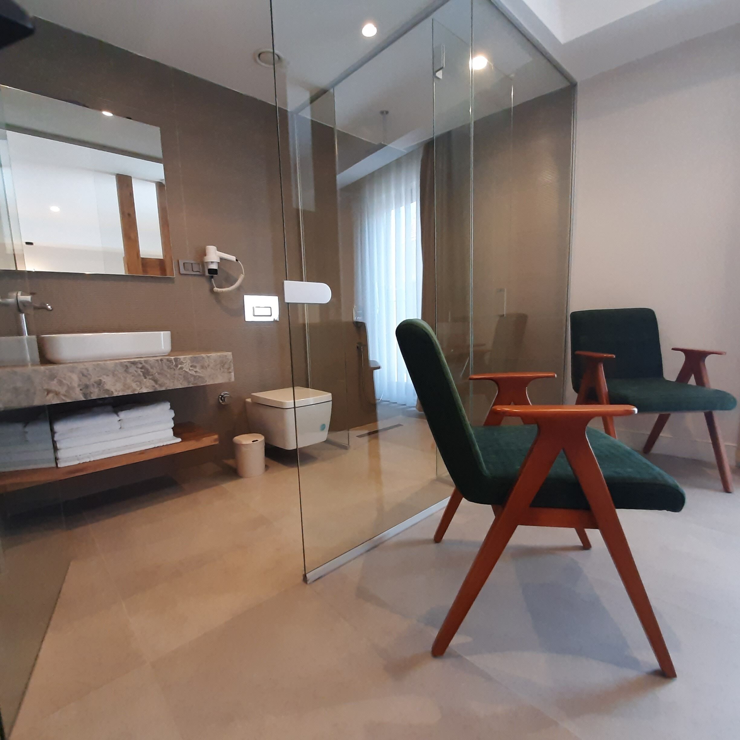 A modern bathroom with a glass partition, a sink, a toilet, and two green chairs.