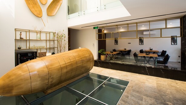A modern interior with a large wooden sculpture, glass flooring, and a reception area with seating and a counter. - Nilus Urla (Urla)