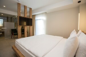 Una habitación de hotel moderna con una cama grande, cabecera de madera, televisor de pantalla plana montado en un panel de madera, un escritorio con silla, y un ventanal con cortinas de gasa.