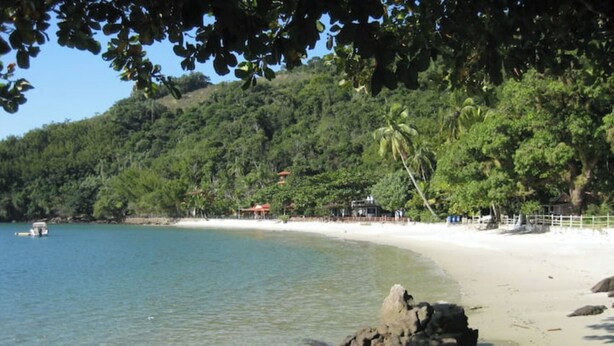 Ein Strand mit klarem Wasser, einem Sandstrand und einer hügeligen Landschaft mit Bäumen im Hintergrund.