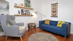 A living room with a grey armchair, a blue sofa, a round wooden table, and a wall-mounted lamp.