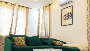 A green sofa with patterned cushions, a white side table, and a framed artwork on the wall.