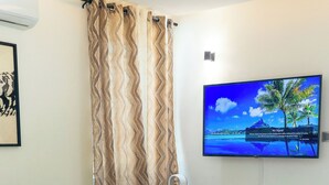 A modern living room with a green sofa, a flat-screen TV mounted on the wall, a white entertainment unit below, and a patterned curtain.