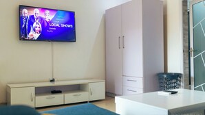 A modern living room with a flat-screen TV displaying local shows, a white cabinet, and a trash bin.