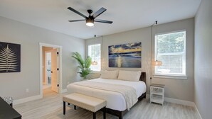 Un dormitorio con cama, ventilador de techo, banco, mesita y ventana con persianas.